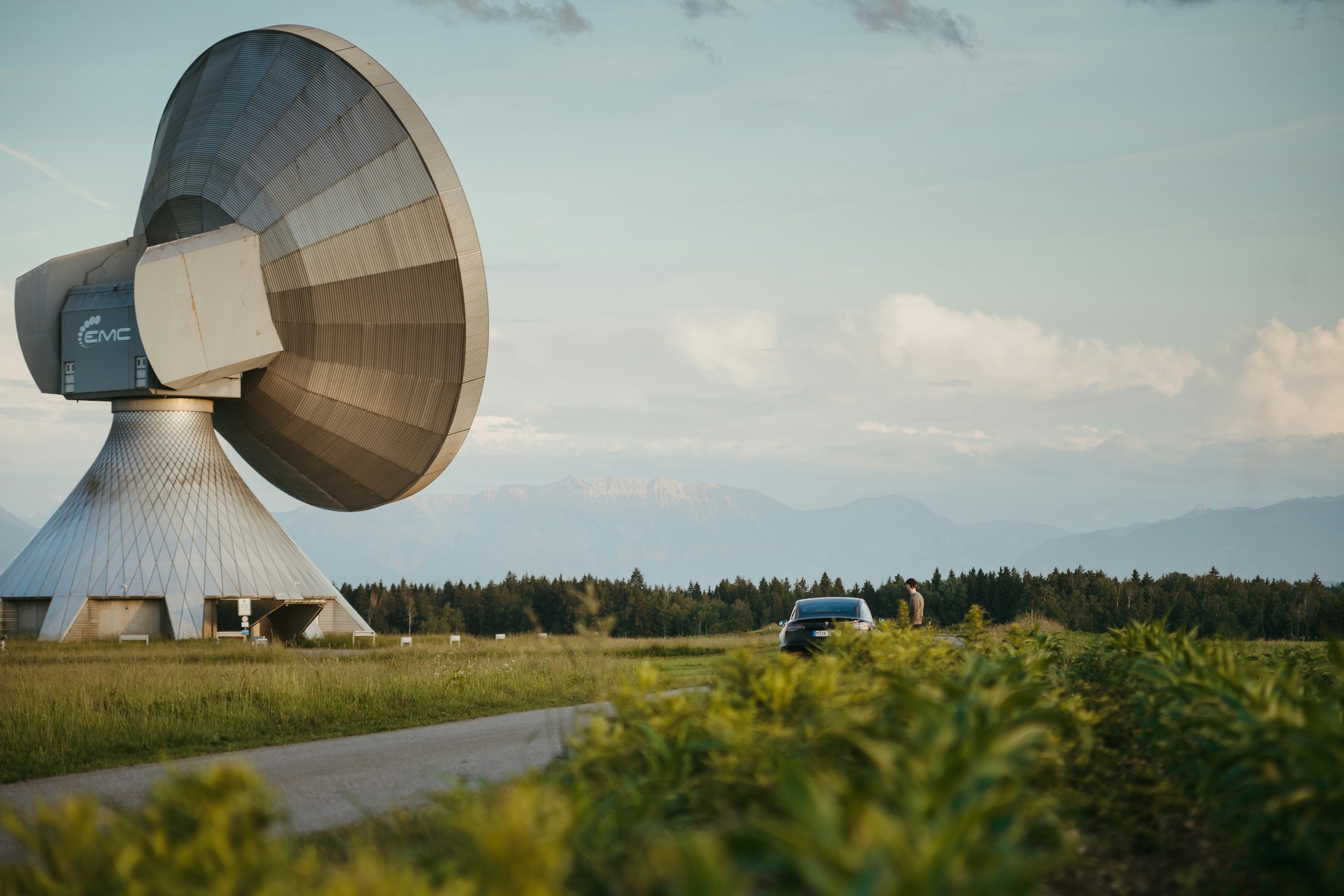 Large satellite communication dish in an open field with mountains in the background and a car.
